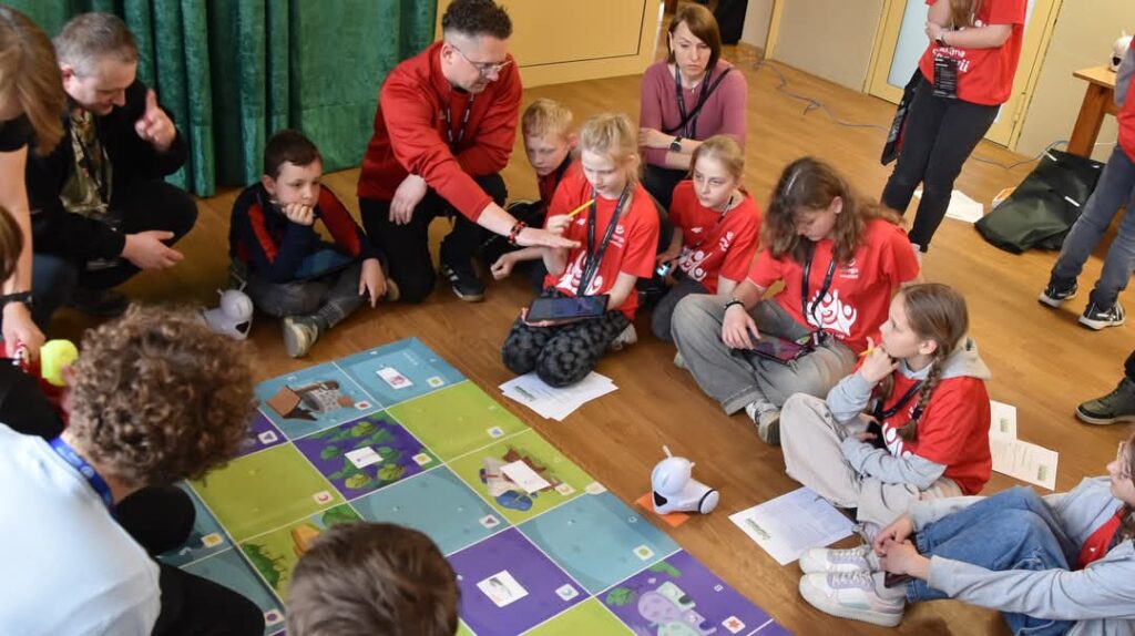 Grupa dzieci i dorosłych siedzi na podłodze w kręgu wokół kolorowej planszy edukacyjnej. Dzieci, ubrane w czerwone koszulki, biorą udział w zajęciach z programowania z wykorzystaniem robota edukacyjnego. Instruktor wskazuje coś na planszy, a dzieci z zainteresowaniem obserwują. Na planszy znajdują się różnokolorowe pola i obrazki, a obok leży biały robot przypominający psa.