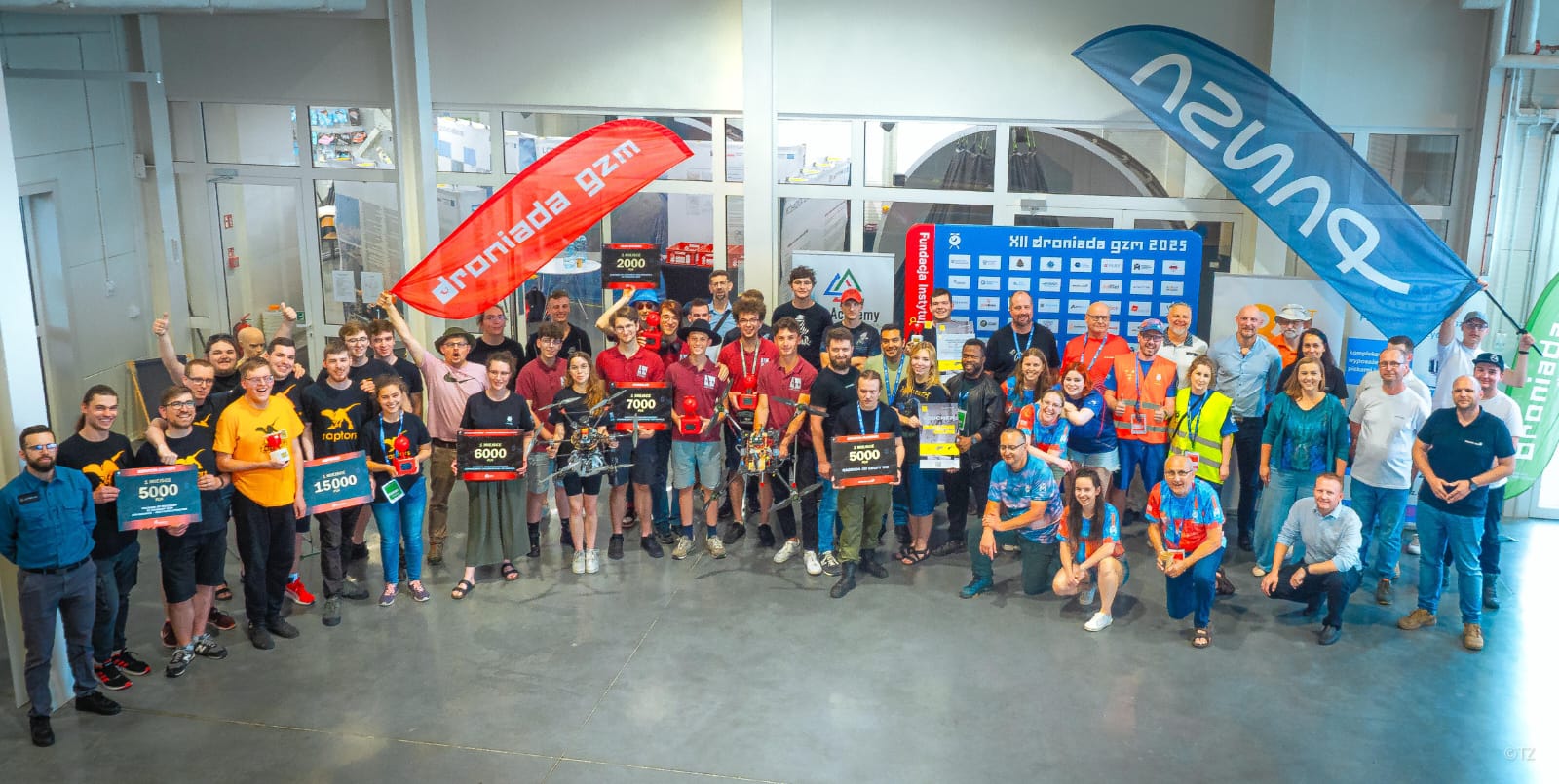 Grupowe zdjęcie uczestników i organizatorów Droniady GZM 2025 w Gliwicach. W centralnej części znajdują się zwycięskie zespoły trzymające duże tablice z kwotami nagród (np. 15 000 zł, 7000 zł, 5000 zł). Uczestnicy pozują z dronami, banerami partnerów wydarzenia i flagami Droniada GZM. W tle widoczna jest tablica wyników i logotypy organizatorów. Zdjęcie wykonane w jasnym, nowoczesnym wnętrzu hali.